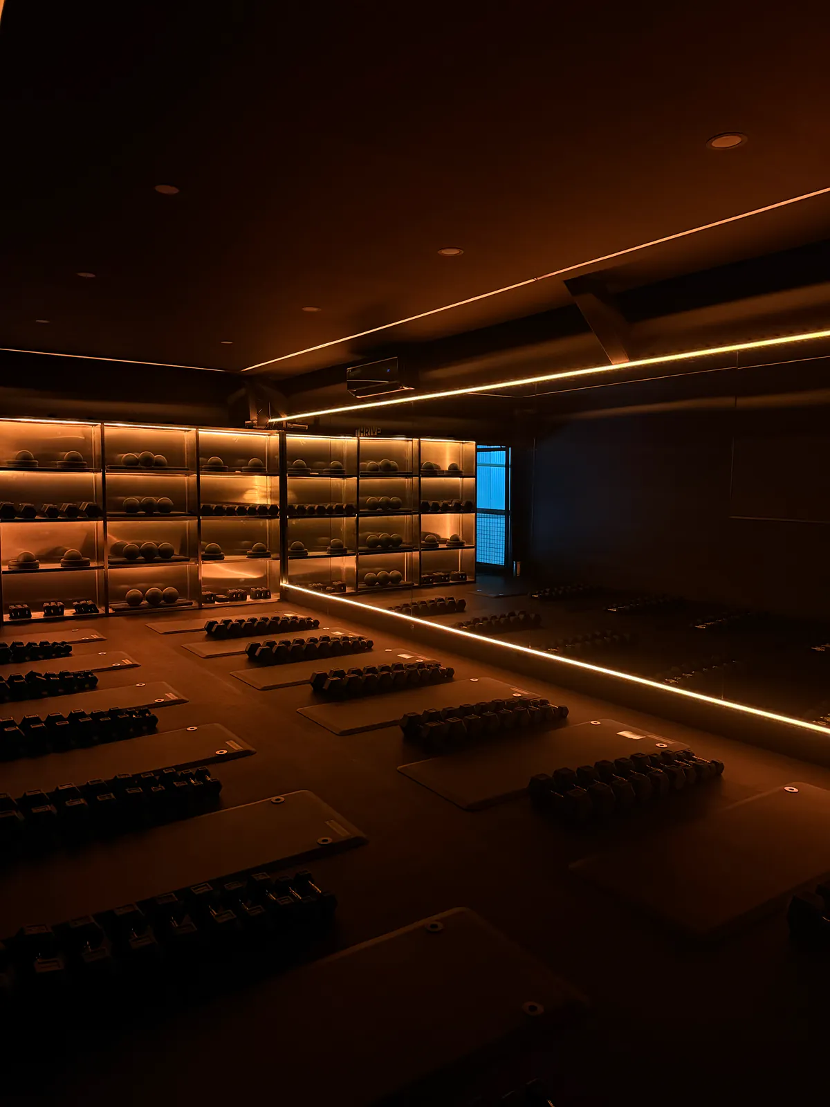The Thr1ve studio floor before a class: mats and dumbbells set out at individual stations under warm light, beside an illuminated wall of weights